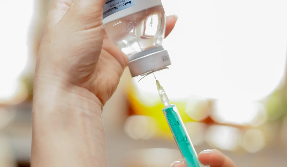 A close-up of a person's hands wearing a white sleeve, holding a clear glass vial upside down while withdrawing liquid into a medical syringe with a green plunger.