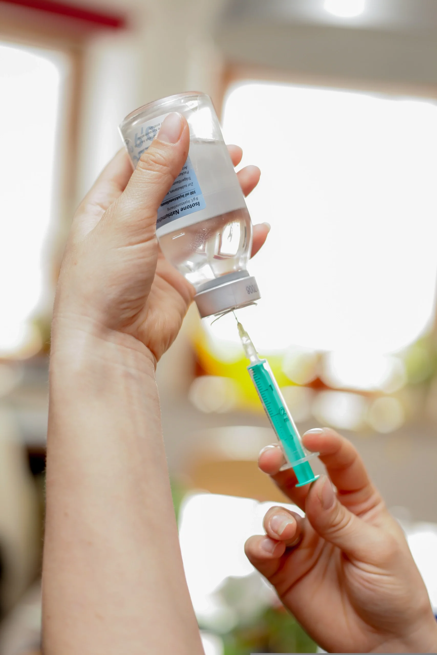 A close-up of a person's hands wearing a white sleeve, holding a clear glass vial upside down while withdrawing liquid into a medical syringe with a green plunger.