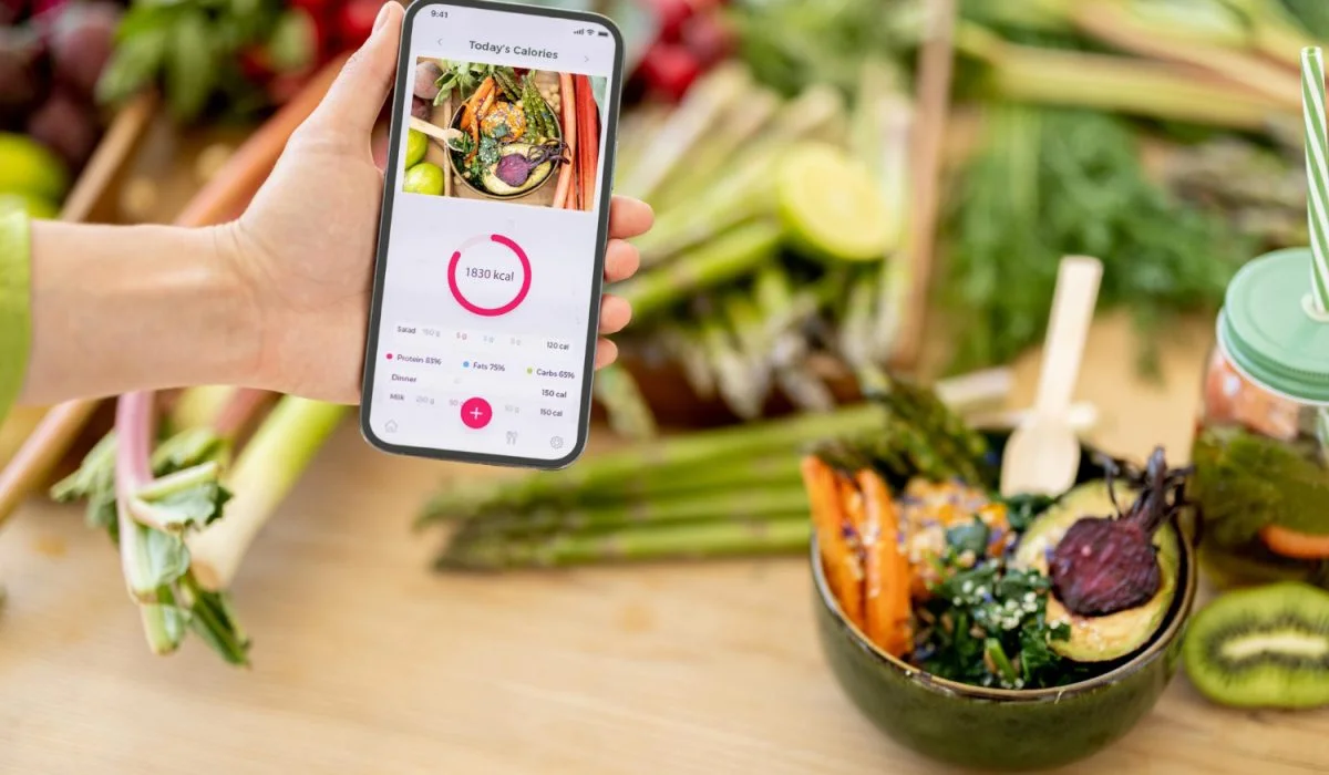 A hand holding a smartphone displaying a calorie-tracking app with a "1830 kcal" progress circle. In the background, a healthy meal bowl with roasted vegetables, avocado, and beets sits on a wooden table surrounded by fresh asparagus and rhubarb.