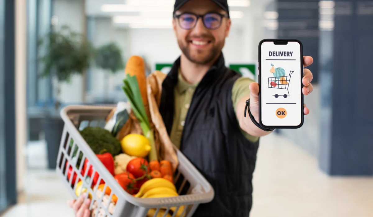 A smiling delivery worker wearing glasses and a cap, holding a plastic basket filled with fresh vegetables and fruits in one hand while showing a smartphone screen with a grocery delivery app interface in the other.