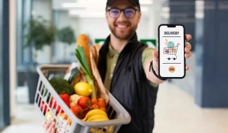 A smiling delivery worker wearing glasses and a cap, holding a plastic basket filled with fresh vegetables and fruits in one hand while showing a smartphone screen with a grocery delivery app interface in the other.