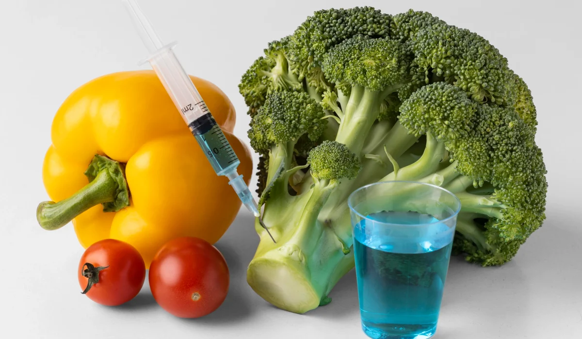 A close-up of a yellow bell pepper, cherry tomatoes, and a head of broccoli on a white surface, with a medical syringe injecting a blue liquid into the broccoli stem.