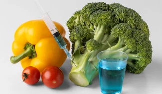 A close-up of a yellow bell pepper, cherry tomatoes, and a head of broccoli on a white surface, with a medical syringe injecting a blue liquid into the broccoli stem.