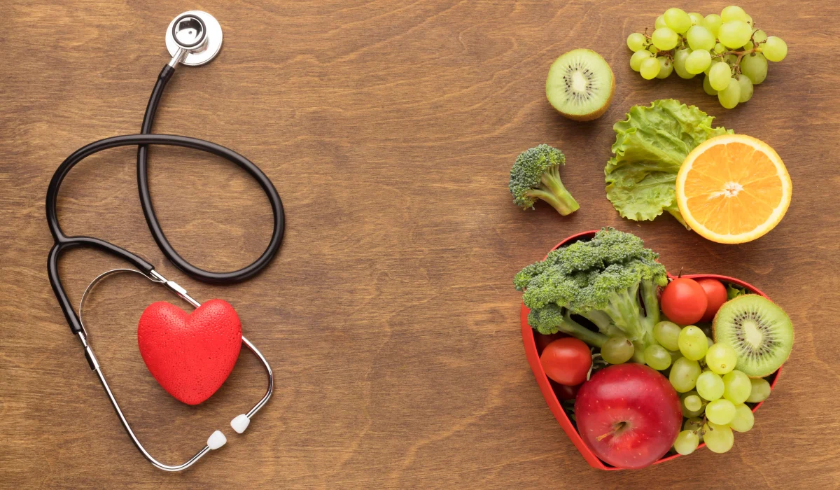 A top-down view of a wooden surface with a stethoscope and a red heart on the left, and a variety of fresh fruits and vegetables—including apples, grapes, and broccoli—arranged on the right.
