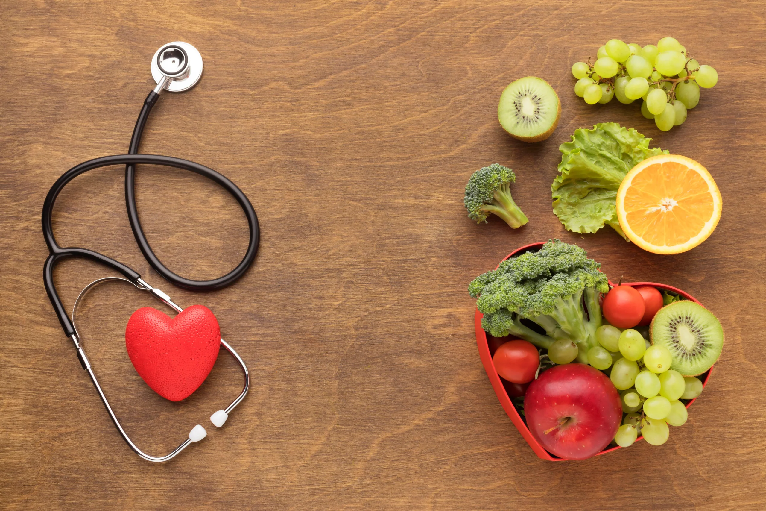 A top-down view of a wooden surface with a stethoscope and a red heart on the left, and a variety of fresh fruits and vegetables—including apples, grapes, and broccoli—arranged on the right.