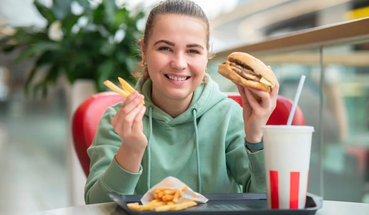 Smiling young woman enjoying a burger, fries, and soda at a fast-food restaurant while tracking her macros with a flexible dieting approach.