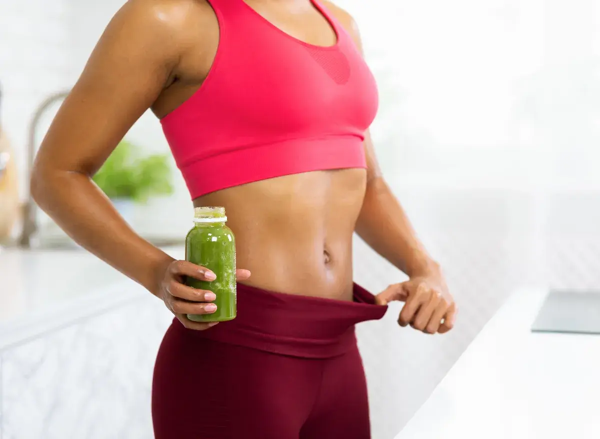 Fit woman in workout clothes holding a green smoothie and pulling her waistband to show weight loss.