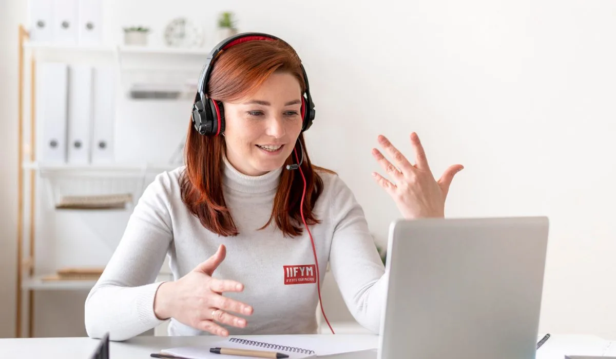 A muscular man in a grey tank top sitting at a wooden table in a bright kitchen, eating a bowl of oatmeal topped with blueberries and bananas. A white banner at the bottom reads "Does carb back loading work?" with the blue IIFYM logo in the top left corner.