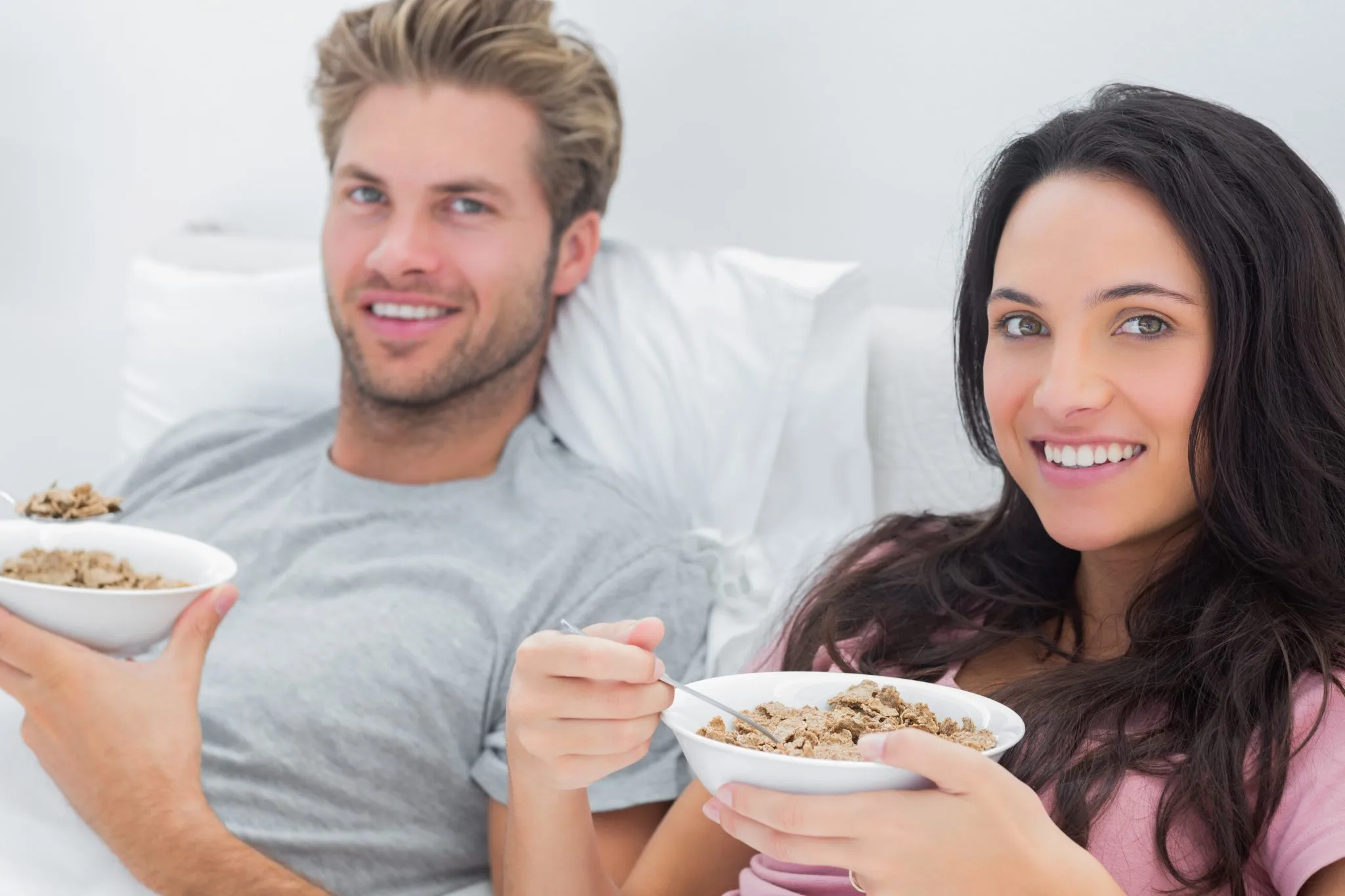 A happy couple, a man and a woman, sitting upright in a white bed and smiling at the camera while holding bowls of bran flake cereal with spoons, enjoying a healthy breakfast in bed.