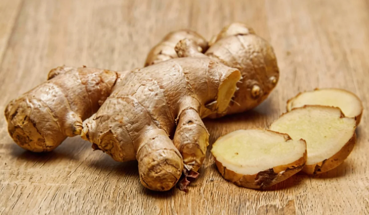 A whole fresh ginger root and three thin slices resting on a light wooden surface.
