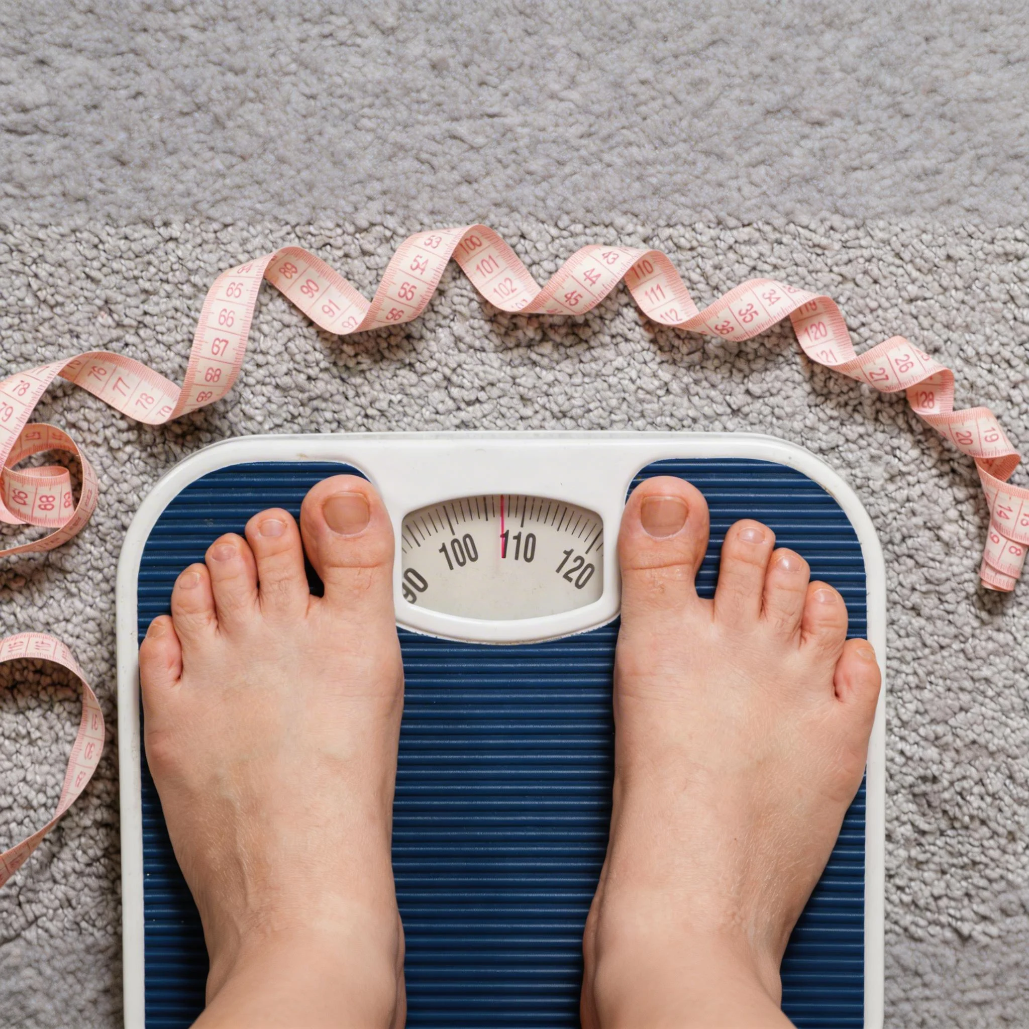 A top-down view of a person's feet standing on a blue and white analog bathroom scale, with a pink measuring tape curled around the scale on a gray carpet.