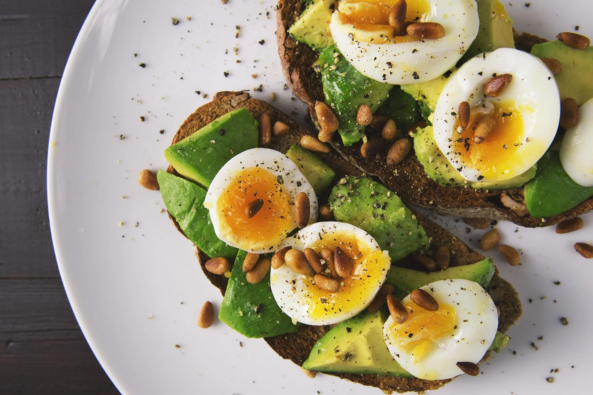 A top-down close-up of two slices of dark whole-grain toast on a white plate, topped with sliced avocado, soft-boiled eggs with runny yolks, toasted pine nuts, and black pepper.