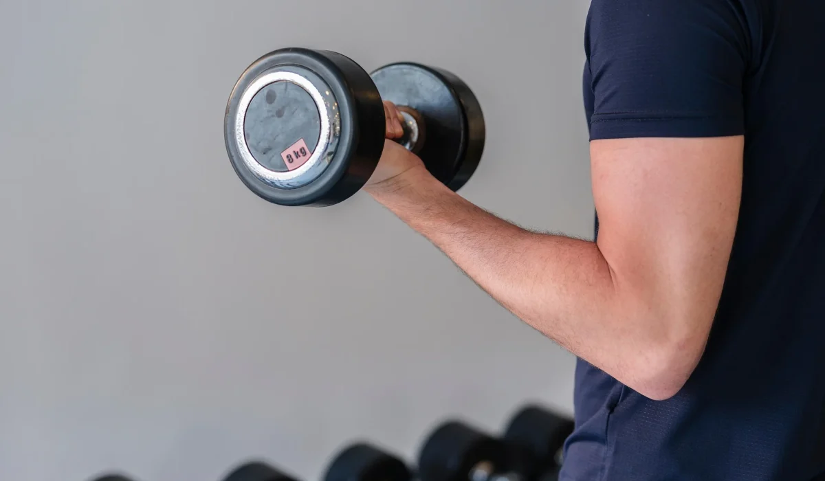A close-up side view of a man in a navy blue t-shirt performing a bicep curl with a black dumbbell in a gym setting, with a rack of weights blurred in the background.