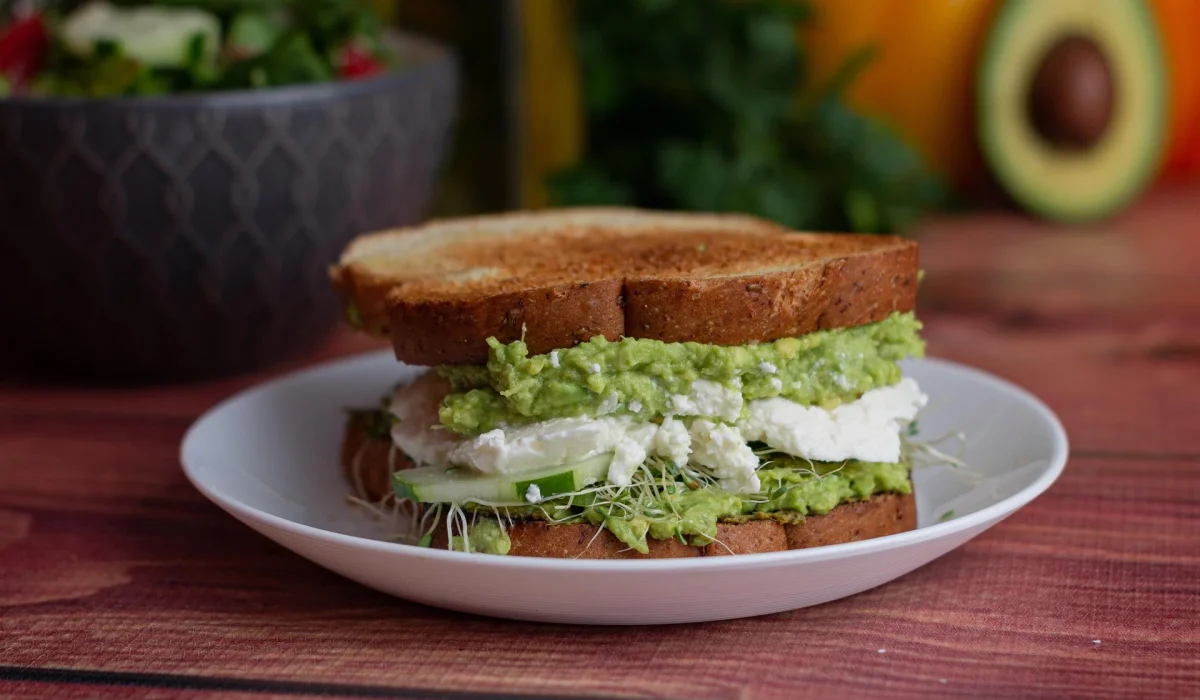 A close-up of a toasted sandwich on a white plate filled with mashed avocado, white cheese, cucumber slices, and fresh sprouts.
