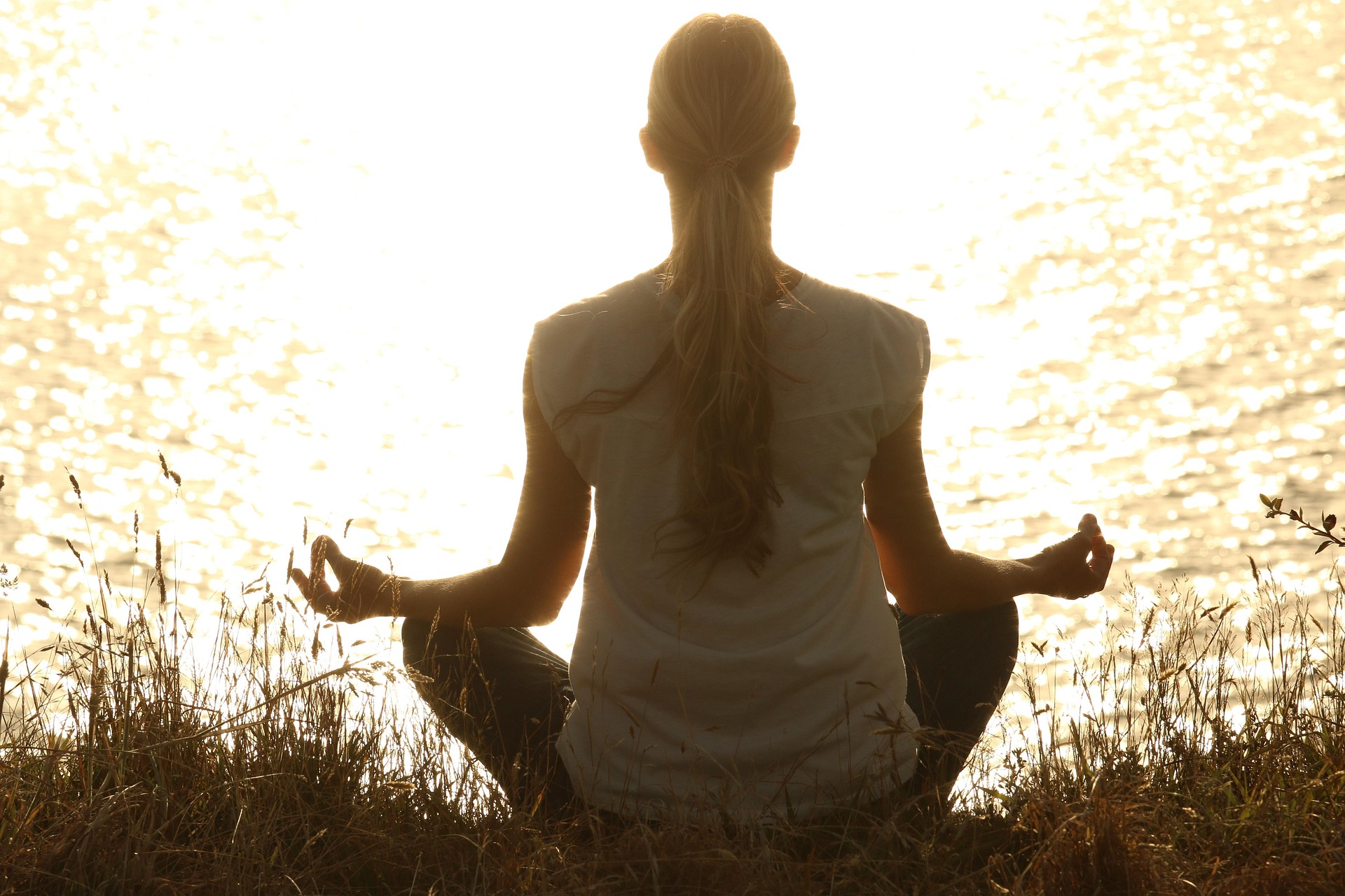 A back view silhouette of a woman sitting in a meditative yoga pose on a grassy shore, facing a bright, shimmering lake at sunset.