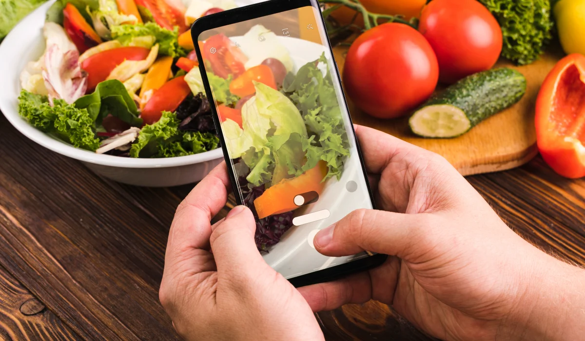 A close-up of a person's hands holding a smartphone to take a photo of a fresh garden salad in a white bowl, with tomatoes and cucumbers visible on a wooden table in the background.