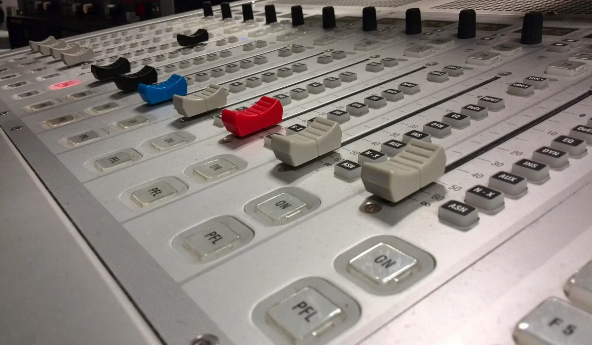 A close-up, angled view of a white professional audio mixing console featuring various faders in black, blue, and red, along with several buttons and knobs.