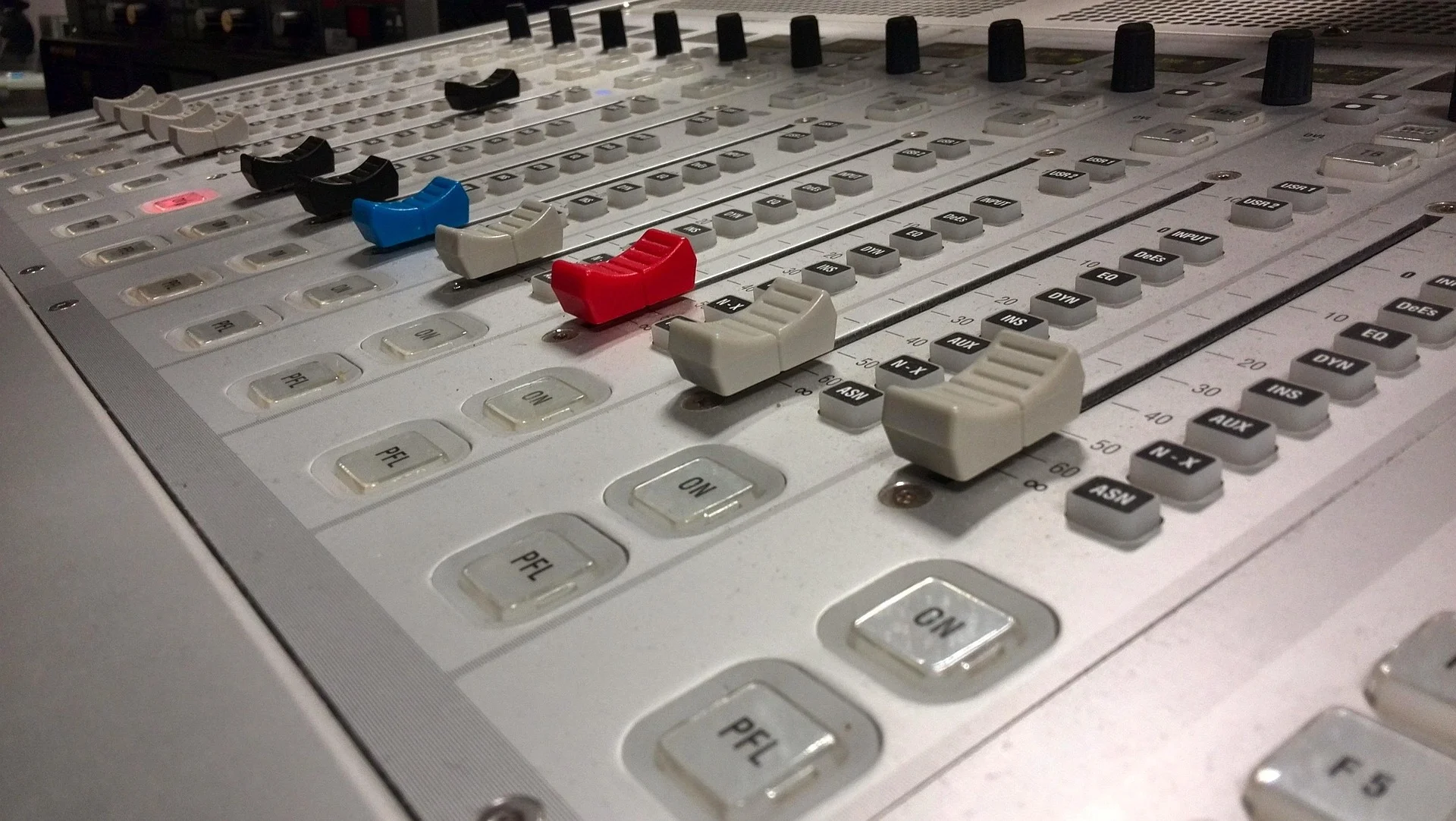 A close-up, angled view of a white professional audio mixing console featuring various faders in black, blue, and red, along with several buttons and knobs.