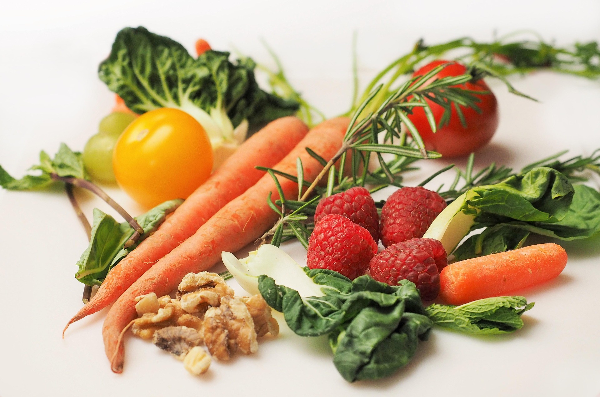 An assortment of fresh whole foods including carrots, raspberries, walnuts, tomatoes, and leafy greens scattered on a white surface.